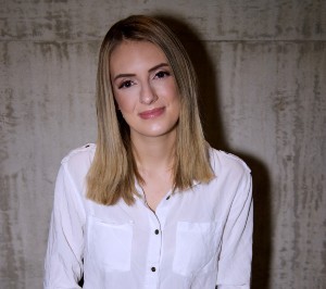 A woman with blonde hair and a white button-up shirt stands in front of a concrete backdrop