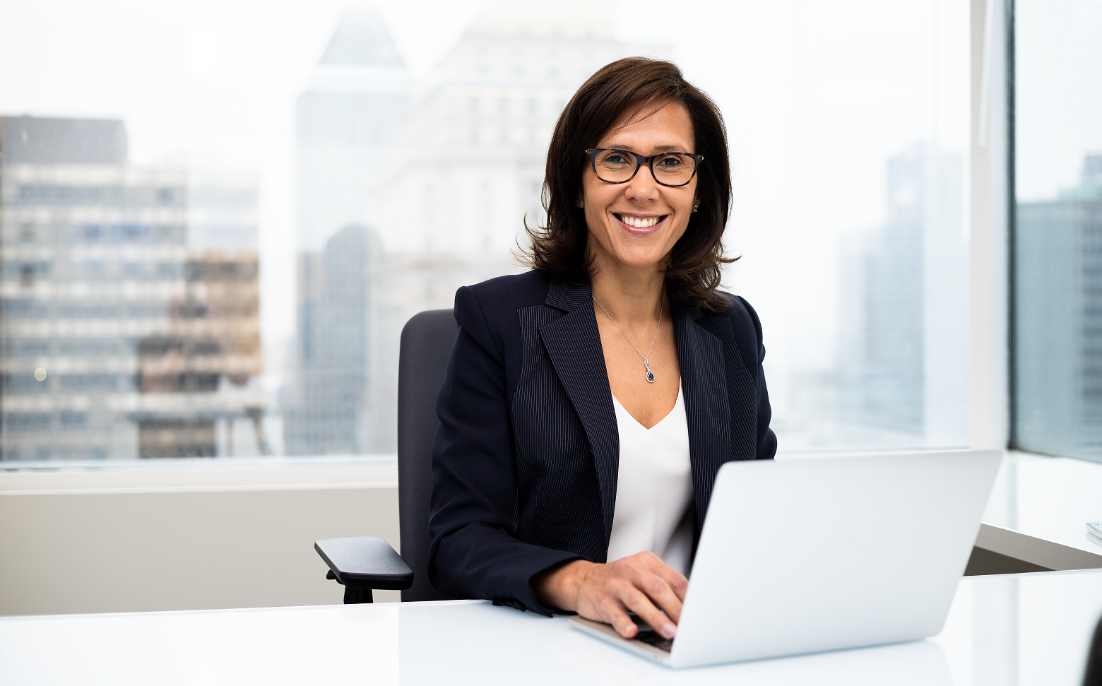 Roula Zaarour sits at an office desk with a laptop in front of large window. She has medium-length, dark brown hair and is wearing a navy-blue blazer with white shirt and dark-framed glasses. 