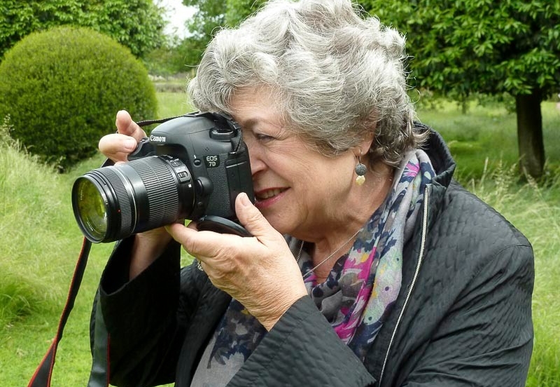 A woman with short light grey hair wears a dark grey jacket and colourful scarf, and looks through a black camera, about to take a photo outdoors