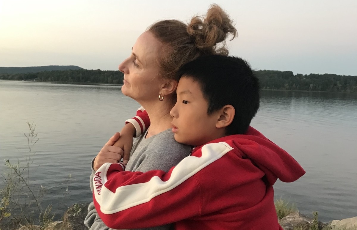 A woman in a grey sweatshirt with light brown hair in a bun looks ahead while her young son, who has short black hair and is wearing a red sweatshirt, hugs her from behind. The pair are standing outside in front of a lake.