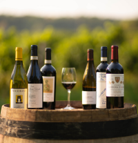 Six bottles and one glass of wine sit atop of a barrel, with vineyard in background