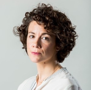 A woman with curly brown hair wears a white shirt with a gold necklace