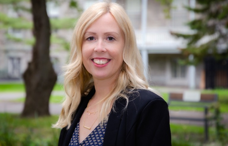 Cherry Marshall A woman with long blonde hair wears a blazer and stands in front of some greenery in the background