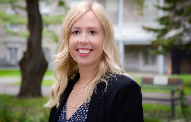 A woman with long blonde hair wears a blazer and stands in front of some greenery in the background