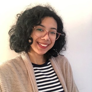 A woman with dark, curly hair wears glasses, a cardigan and a striped shirt