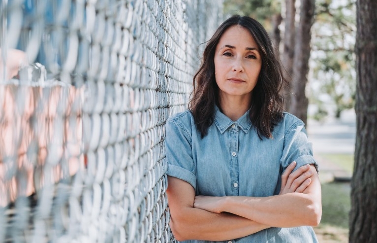 A woman with long dark hair wears a button up shirt and leans against a wired fence