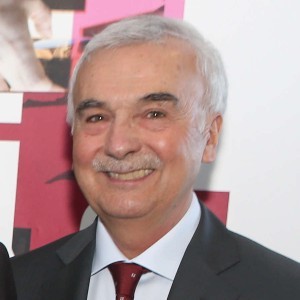 A man with grey hair and mustache wears a dark suit and tie