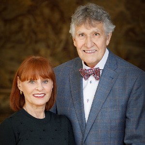 A woman with red hair sits next to a man who is standing and wearing a suit and bow tie