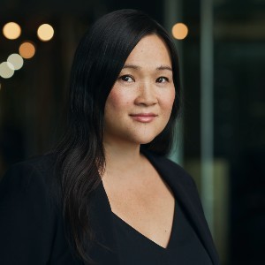 A woman with long black hair wears black clothing in front of a darkened background with some lights