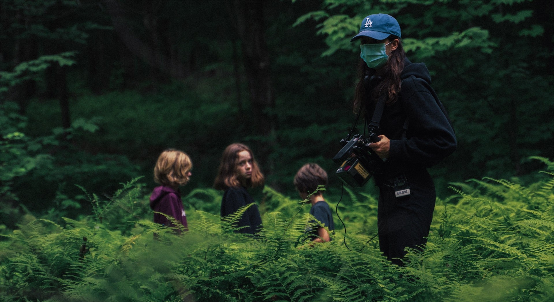 Salomé Villeneuve stands in a field of tall green grass wearing a baseball cap and a surgical mask. She holds a film camera in her hands and to her left are three small children