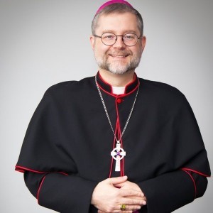 A bishop with a grey beard wears a robe-like vestment with a large cross around his neck