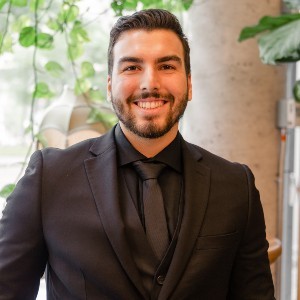 A man with short dark hair wears a black suit and tie