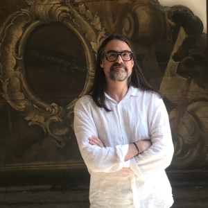 A man with long dark hair and a greying goatee stands with his arms crossed