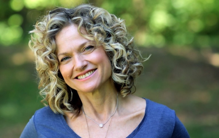 A woman with curly hair stands in front of greenery