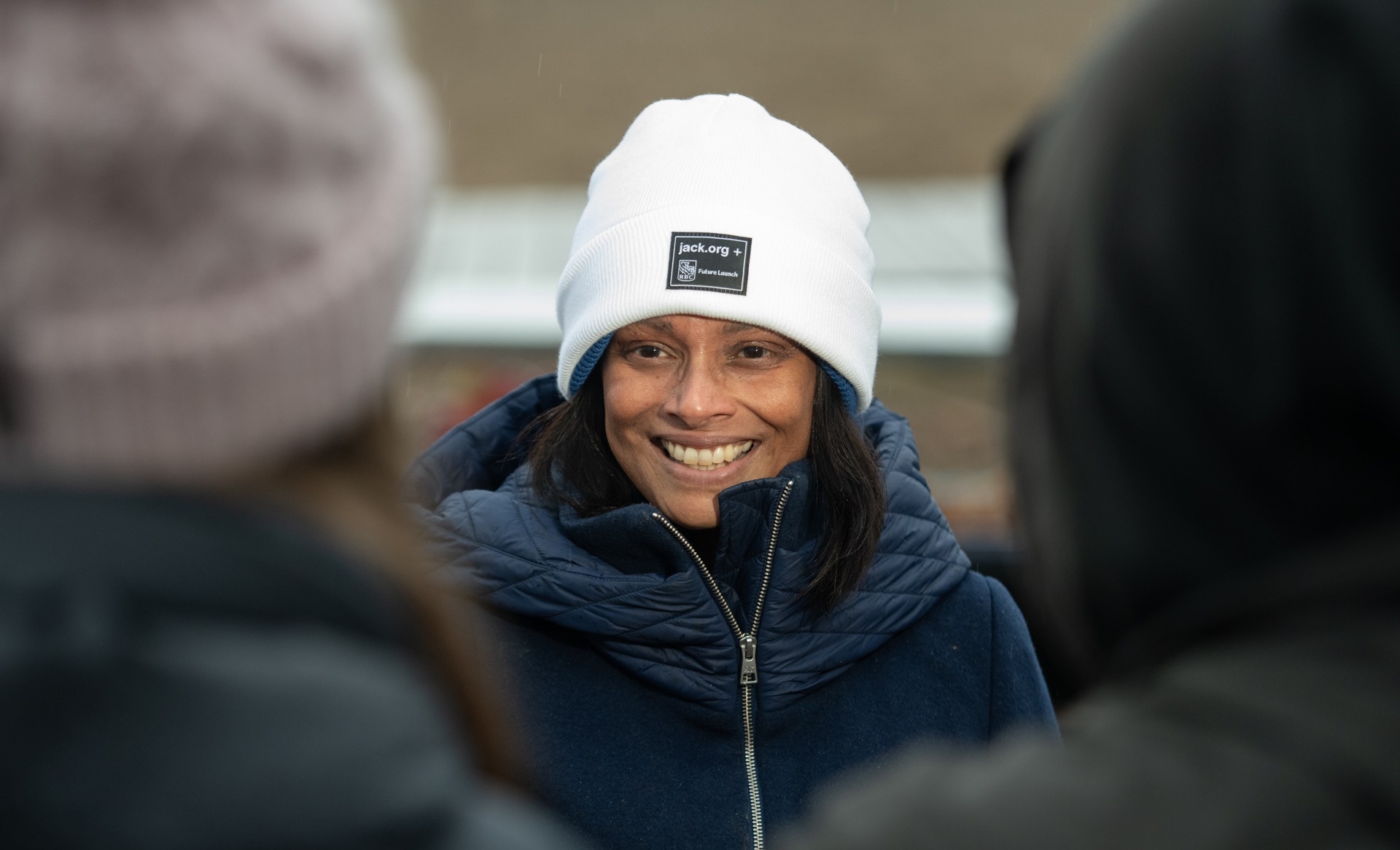A woman is standing outdoors, smiling. She has black hair and is wearing a white tuque and navy blue winter jacket.
