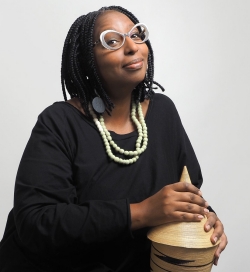 Woman with glasses wearing black dress and beaded necklace, holding small drum