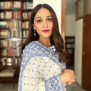 A woman with long brown hair wears a saree with a bookcase in the backgroun