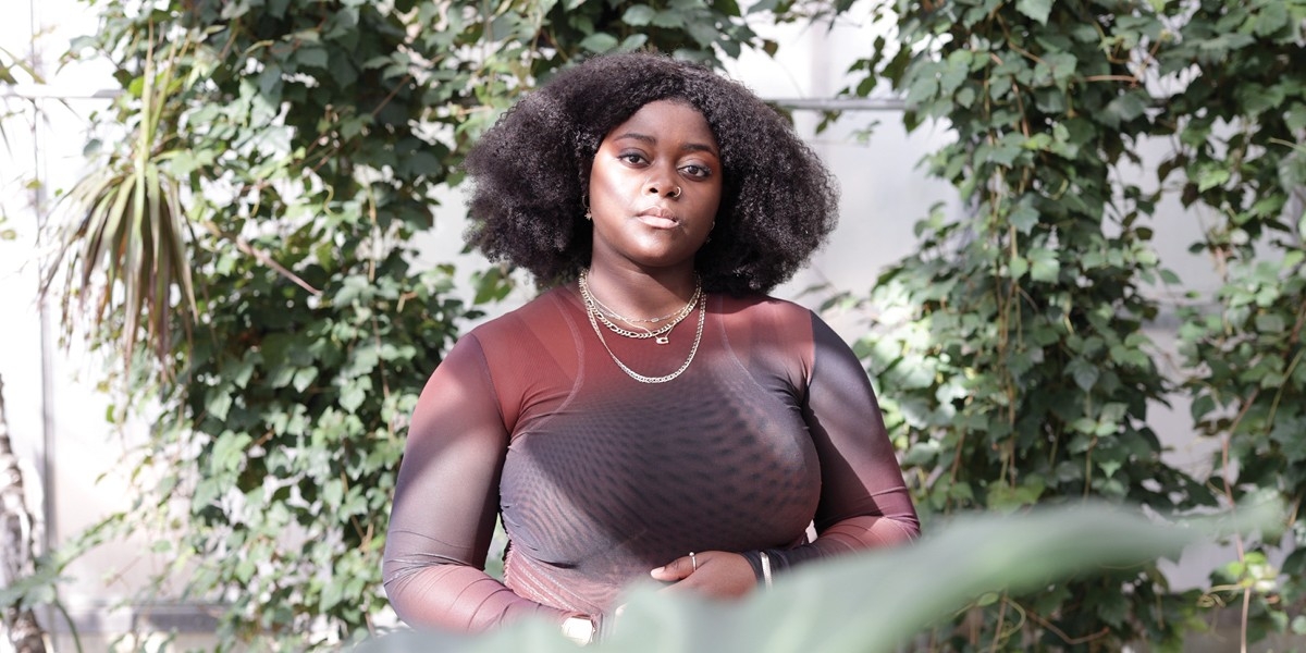 Woman wearing brown top, gold nose ring and chain necklaces standing among plants in Concordia's greenhouse