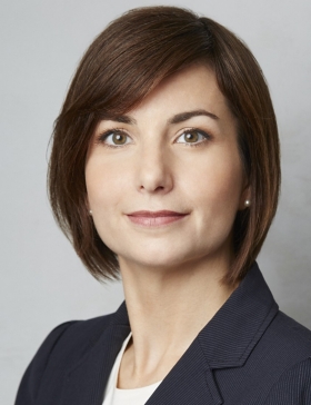 A woman with brown hair wears a blazer in front of a grey background