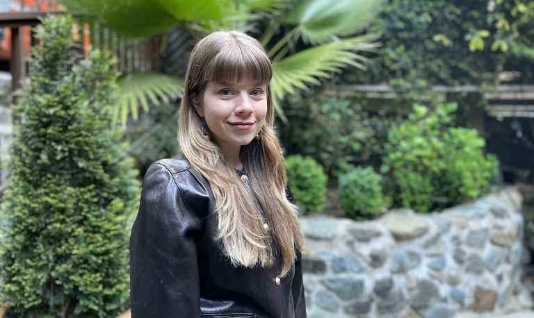 A woman with long blonde hair wears a black leather jacket in front of foliage