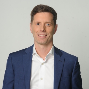 A man with short brown hair wears a navy blue blazer and white shirt