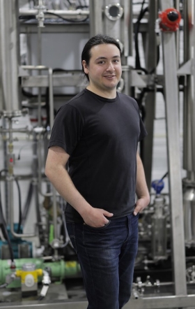 A young man wearing a black T-shirt and jeans stands in front to of machinery in a distillery.