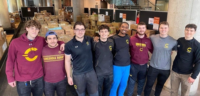 Members of the Men’s Rugby Team stand in front of rows of cardboard boxes filled with books