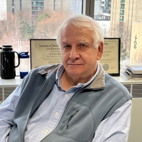 A man with with hairs, wearing a blue shirt and grey fleece vest sits at his desk in an office with a city skyline visible behind him through the window