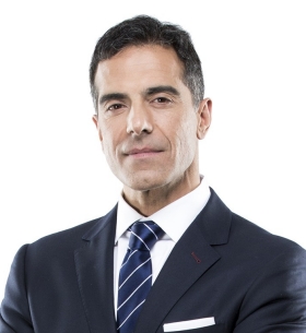 Against a white background, a head and shoulders shot of a man with short dark hair, wearing a dark blue suit, white shirt and blue tie with white stripes