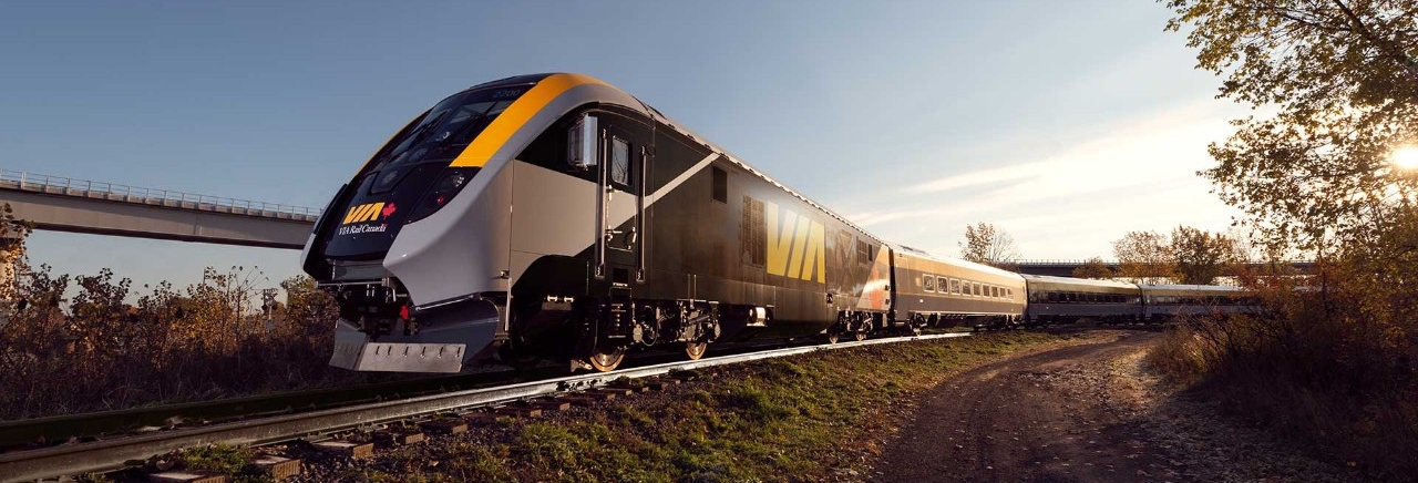 A VIA Rail train locomotive travels along tracks that are surrounded by greenery with a blue sky above.