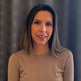 A woman with long straight brown hair wearing a beige thin-knit sweater poses in front of a blue curtain background