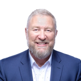 A man with short white hair and a trimmed beard wears a white shirt and a blue blazer 