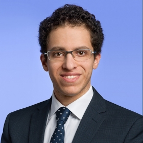 A man with glasses and dark curly hair wears a white shirt, blue tie and dark suit jacket.