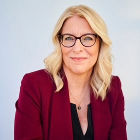 A woman with shoulder-length blonde hair and dark frame glasses wears a black blouse and a red blazer