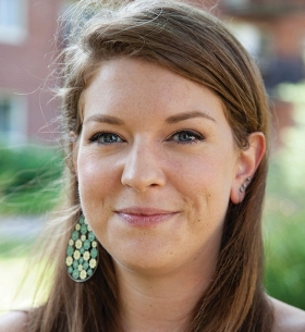 A young woman with long brown hair stands outside with greenery behind her.