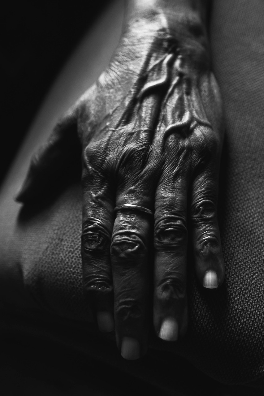 A black and white close-up of an old woman's hand; on her middle finger is a ring