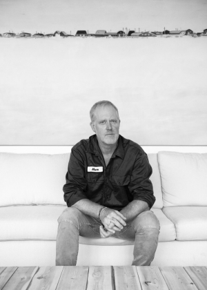 Black and white portrait of a man sitting on a white sofa with large photograph in the background. He is wearing a dark button-down shirt and jeans.