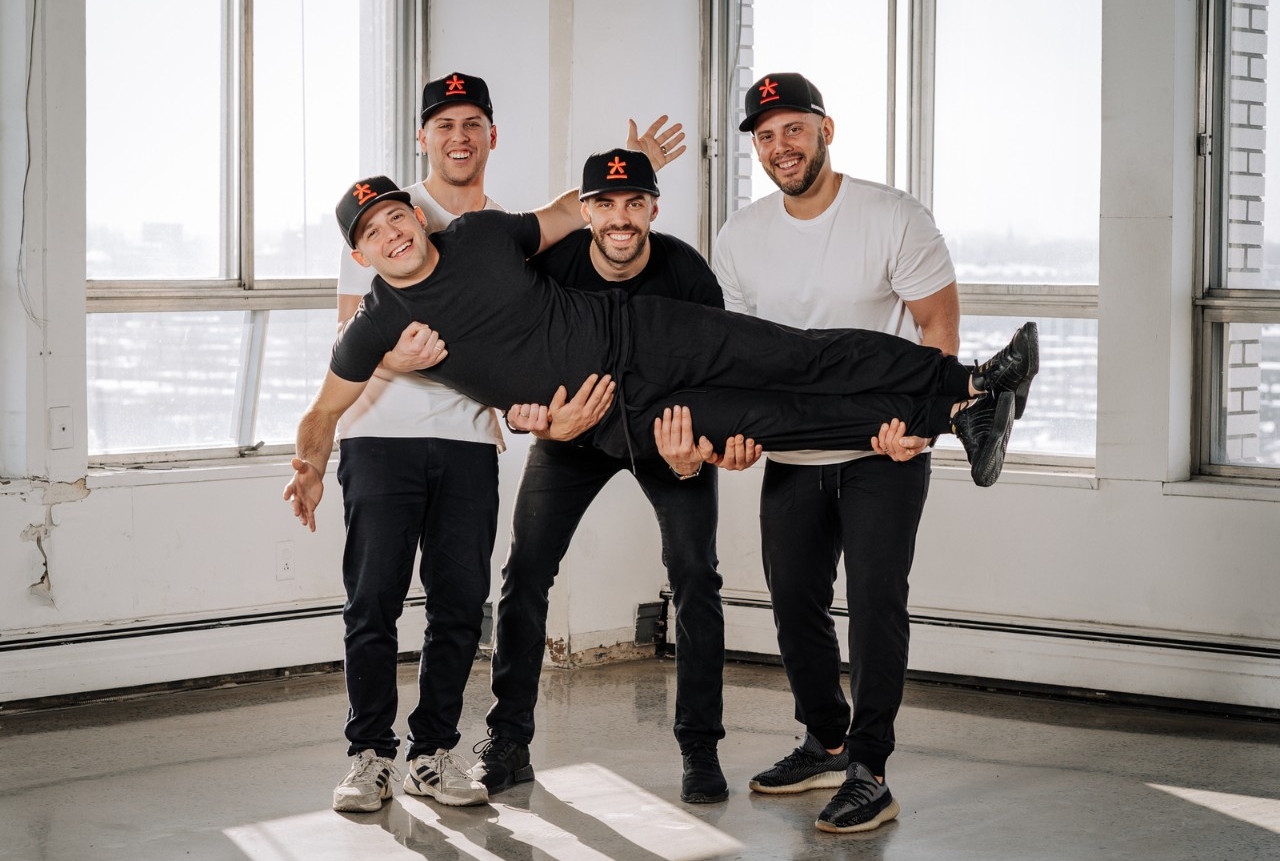 Manmade founders Three men stand next next to each other as they hold a fourth man laying horizontally. They are all smiling and wearing black baseball caps with an orange logo.