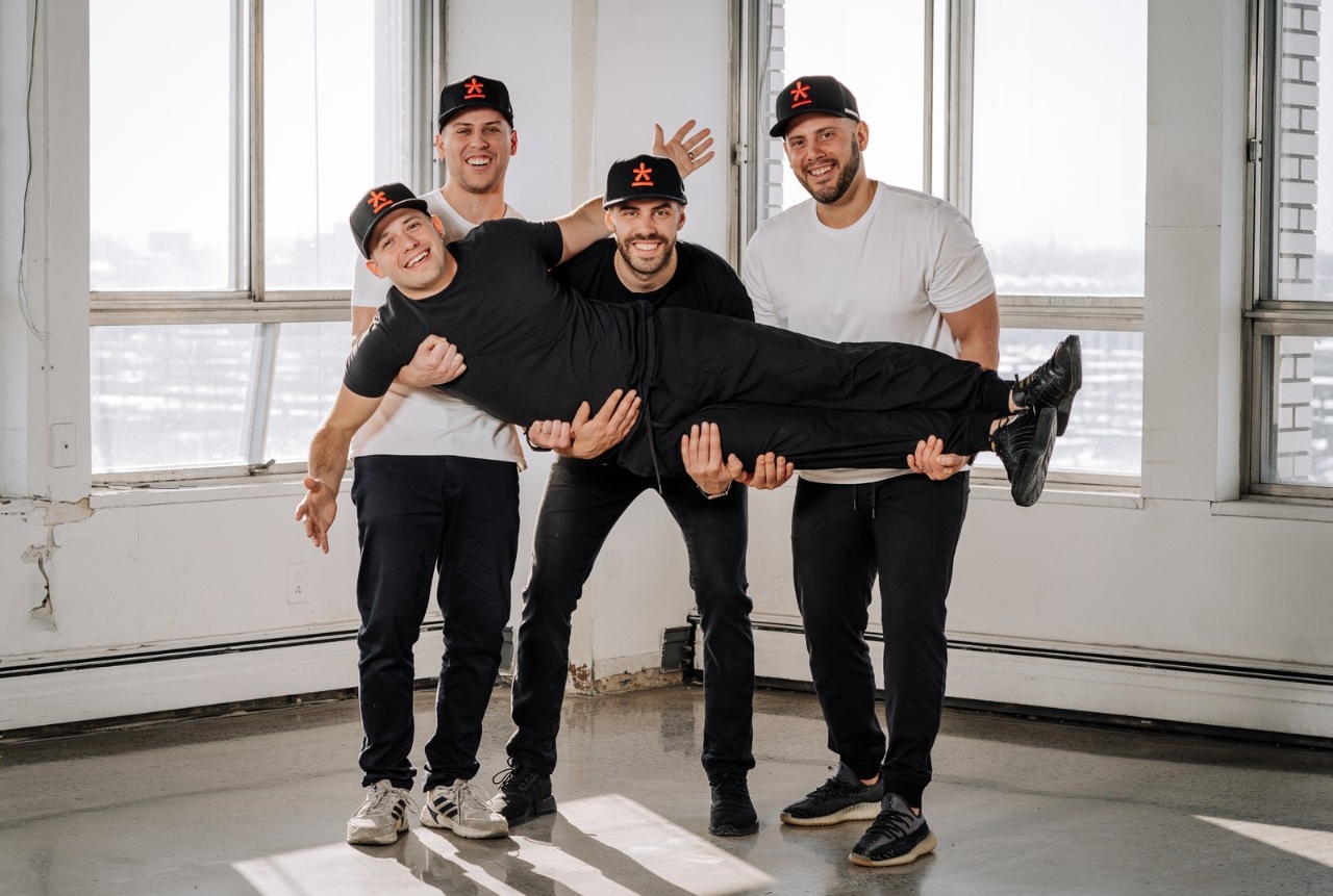 Three men stand next next to each other as they hold a fourth man laying horizontally. They are all smiling and wearing black baseball caps with an orange logo.