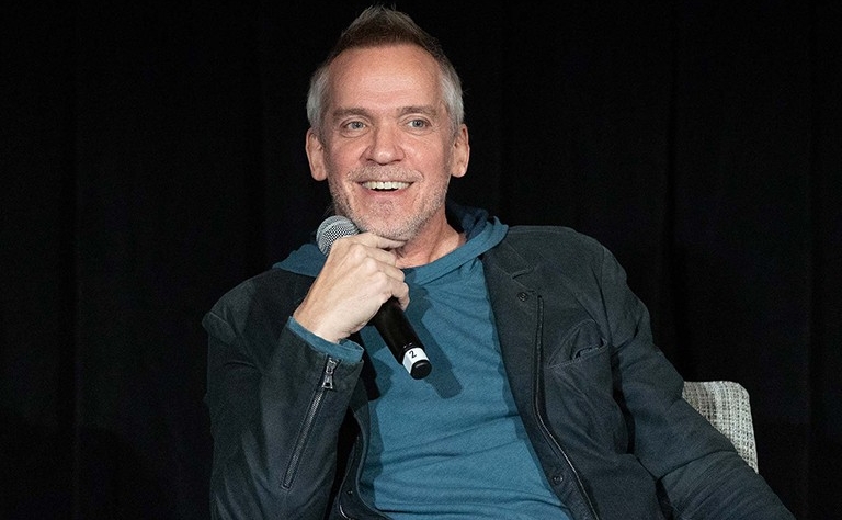 The late Jean-Marc Vallée is pictured sitting down, smiling while holding a mic near his face. He is wearing a blue hoodie under a black jacket.