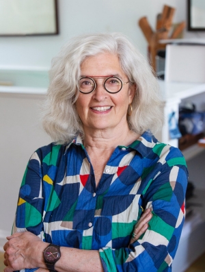 A woman with white hair and black circle-framed glasses sits smiling with her arms crossed. She is sitting in an office and is wearing a colourful button-down shirt