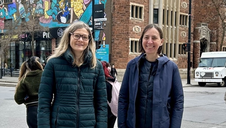 Two women, one with blonde hair and glasses and another with short brown hair, stand next to each other smiling on a Montreal street.