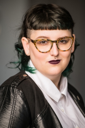 Portrait of a person with short dark hair with green tips wears dark purple lipstick and is wearing multicoloured glasses and a white-collard shirt with a leather black jacket.