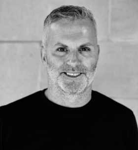 A black and white portrait of a man with short cropped grey hair. He is wearing a black T-shirt and is standing in front of a brick wall.