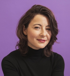 A headshot of a woman with dark wavy hair in a black turtleneck against a deep purple background