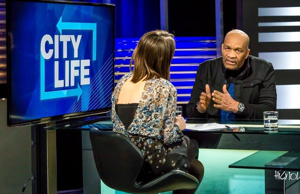 Man wearing black jacket and blue top, sitting at new desk across from newscaster, with TV screen in background