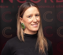 A woman with light brown hair stands is wearing a black turtleneck and green beaded earrings