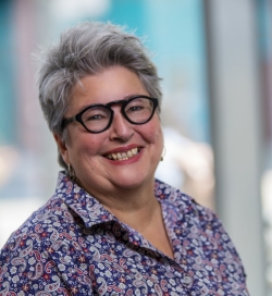 Portrait of a smiling woman with short grey hair wearing black-framed glasses and a collard shirt with a flower pattern.