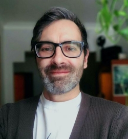 Portrait of a man wearing a dark grey cardican with a light t-shirt beneath. He has a short, grey-brown beard and is wearing black-framed glasses