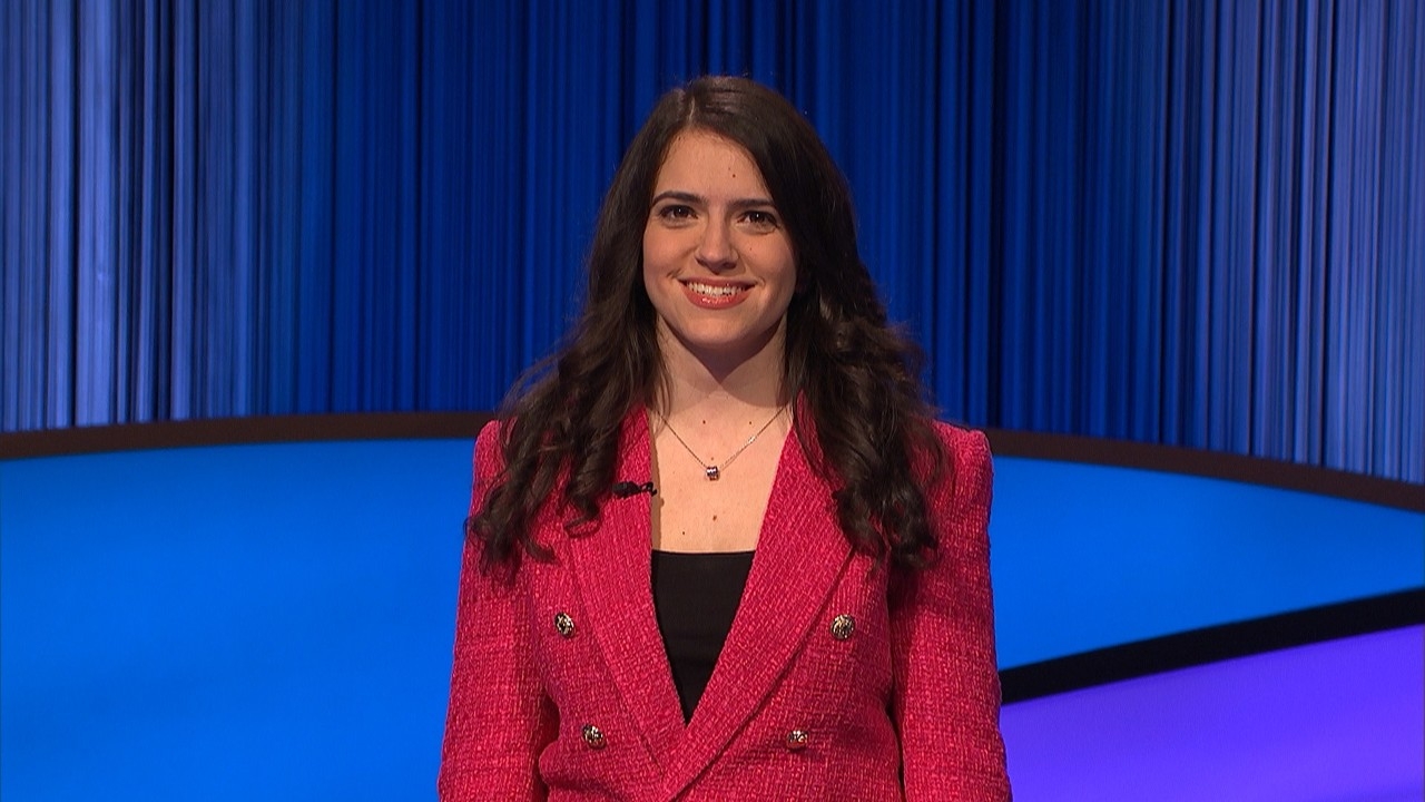 Woman with long, brown hair stands on a blue stage with blue curtains in background